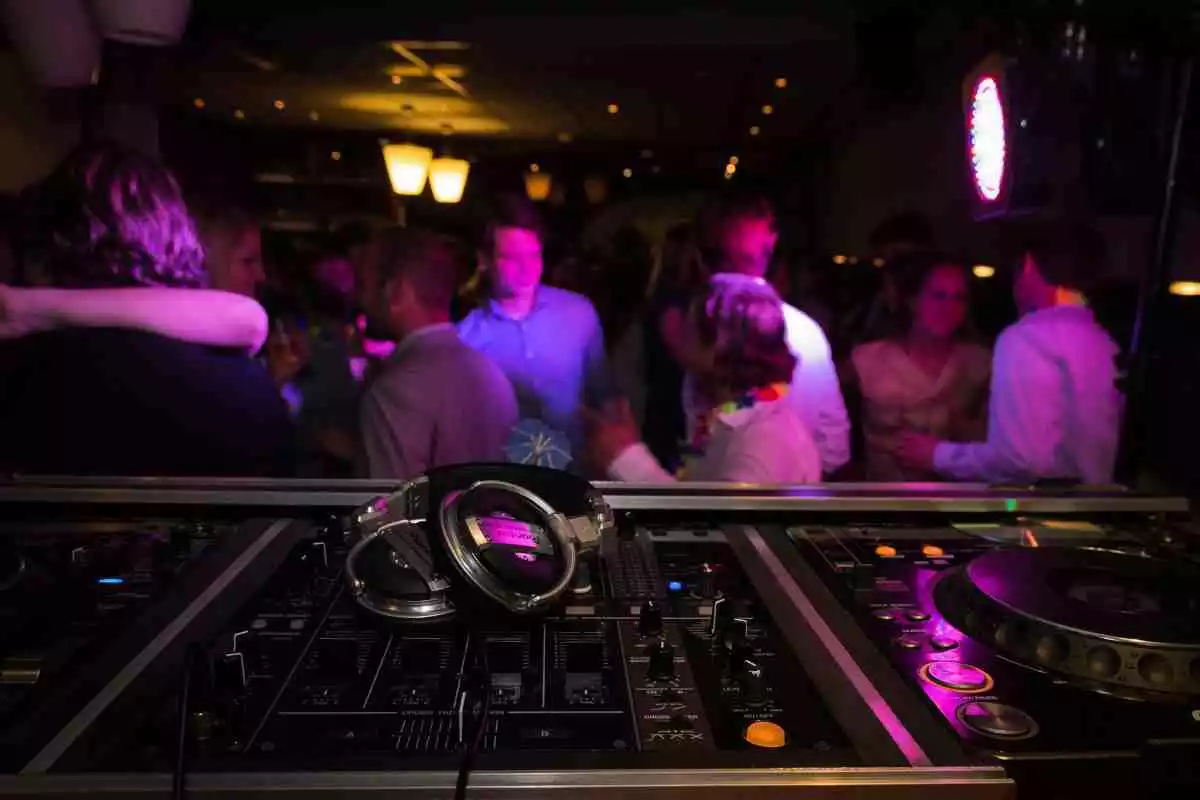 Diverses persones ballen en una festa, amb la taula de mescles i uns auriculars en primer pla