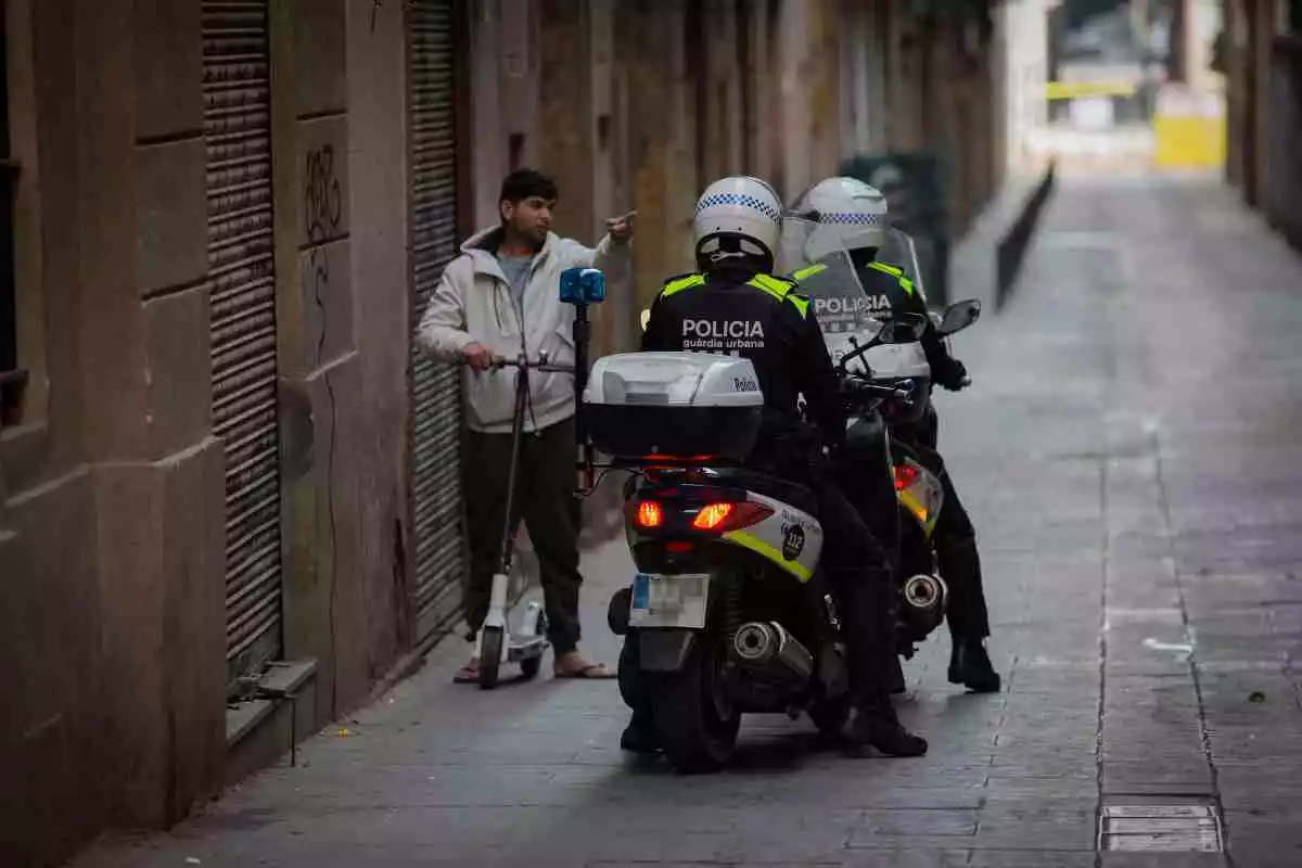 Dos agents de la Guàrdia Urbana de Barcelona, en moto, parlen amb un vianant que no porta mascareta
