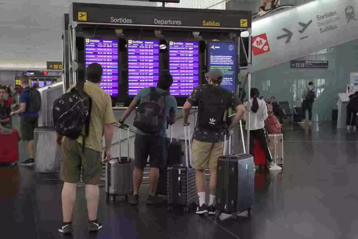 Pla general de tres passatgers de l'aeroport del Prat d'esquenes mirant un panell amb la informació de les sortides de la terminal 2