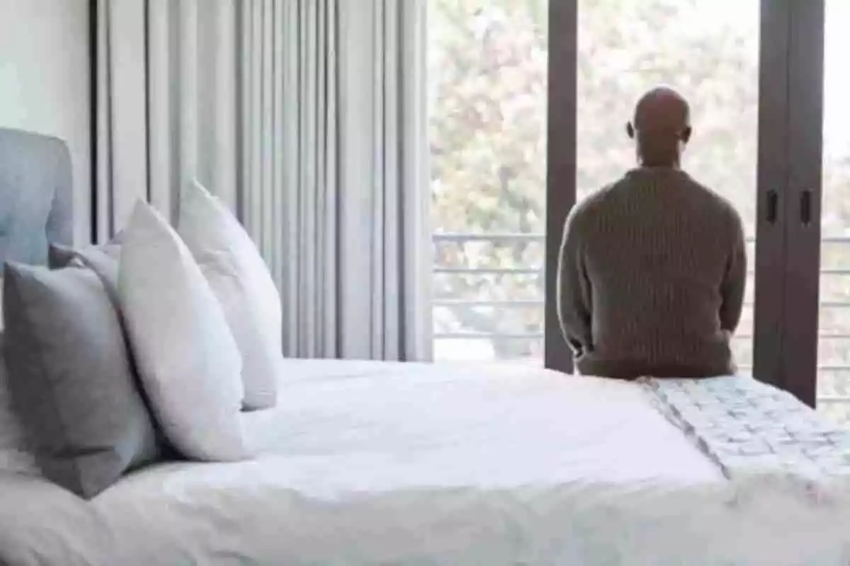 Un home assegut damunt del llit i mirant per la finestra