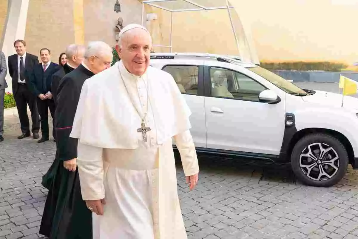 El Papa Francesc en una compareixença pública, vestit de blanc i davant d'un cotxe també blanc, amb gent darrere