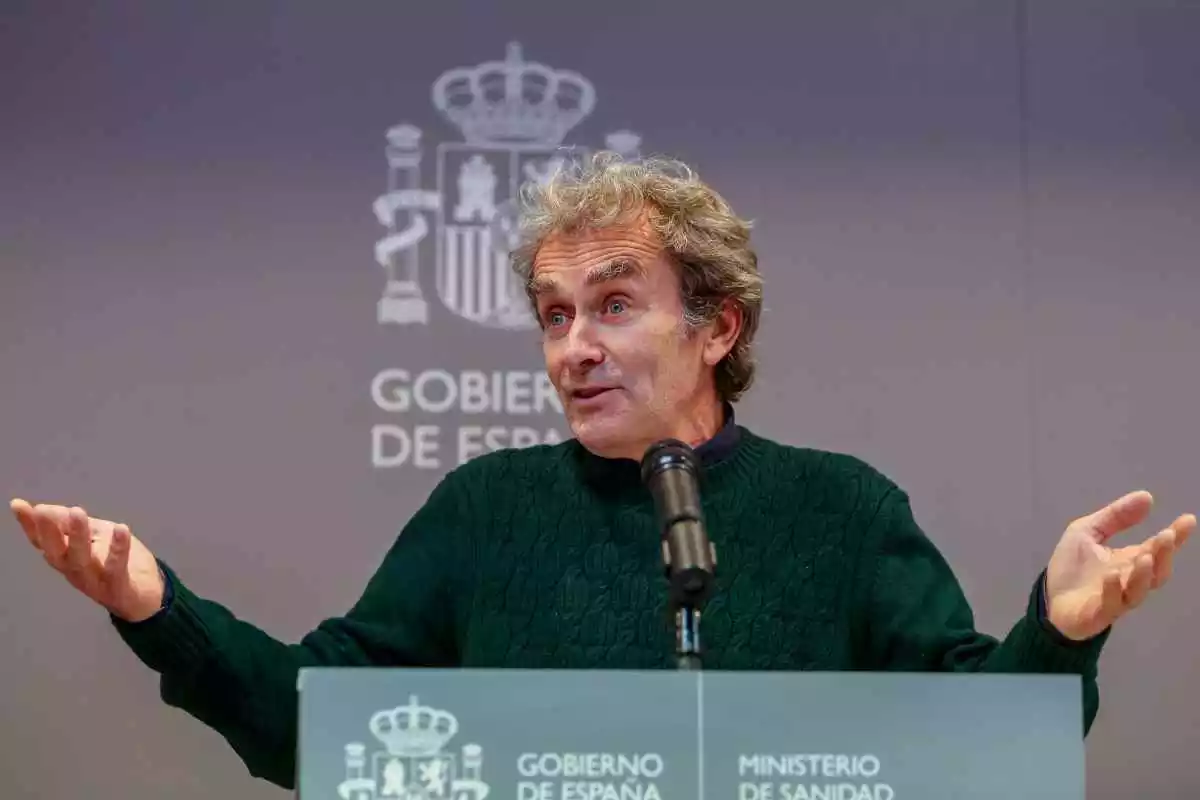 Fernando Simón en roda de premsa aixecant les mans amb cara estranyada i l'escut espanyol darrere