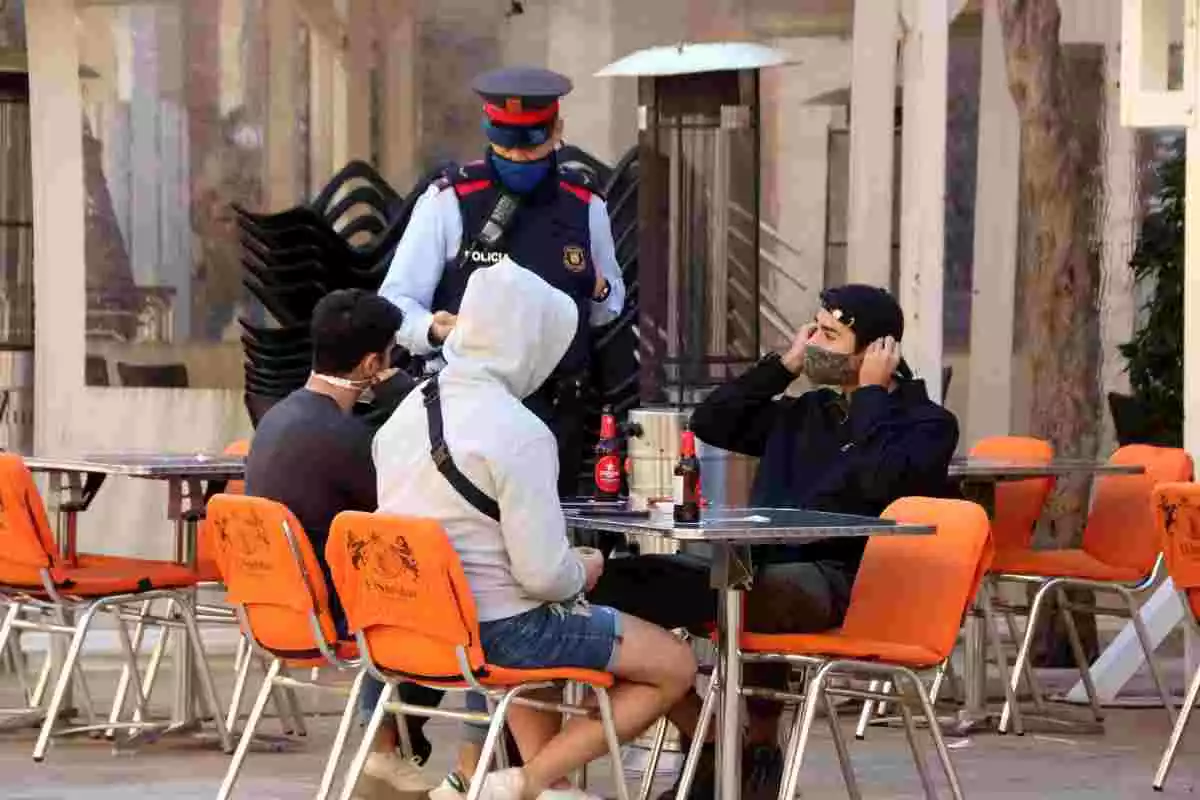 Un agent dels Mossos d'Esquadra parlant amb tres joves asseguts en una terrassa, tots ells amb mascareta
