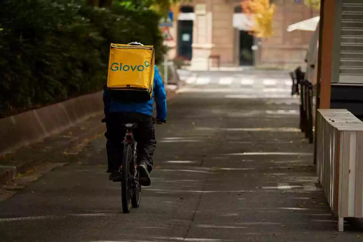 Imatge d'un repartidor de Glovo, d'esquenes, muntant en bicicleta,