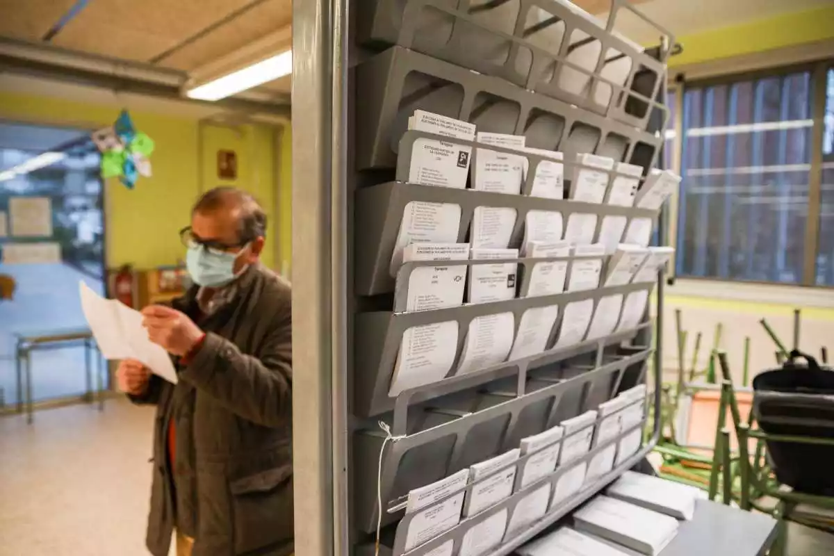 Imatge de paperetes electorals en un col·legi electoral, amb un home en segon pla preparant el seu vot