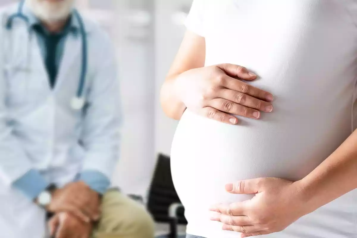 Una dona embarassada acariciant la seva panxa i un doctor assegut de fons