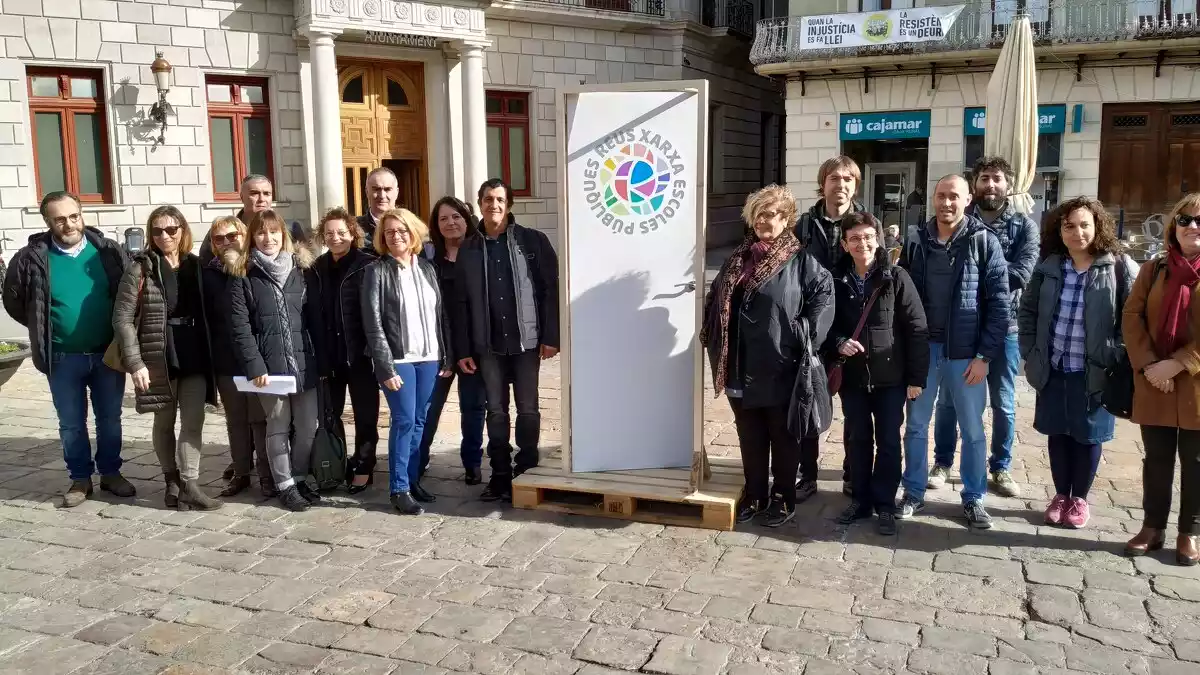 Foto de família dels directors i directores de les escoles públiques de Reus a la plaça del Mercadal, acompanyats del regidor i amb la porta amb el logotip de la Xarxa al mig