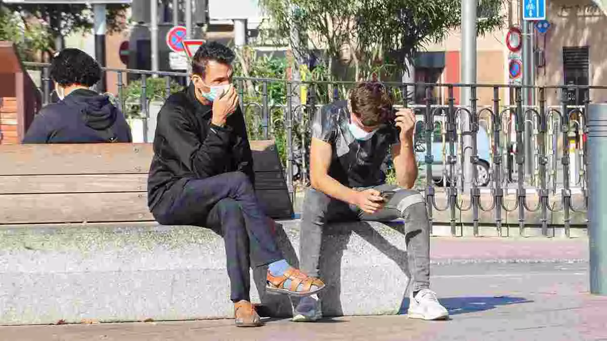 Pla curt de dos joves al carrer amb mascareta asseguts en un banc