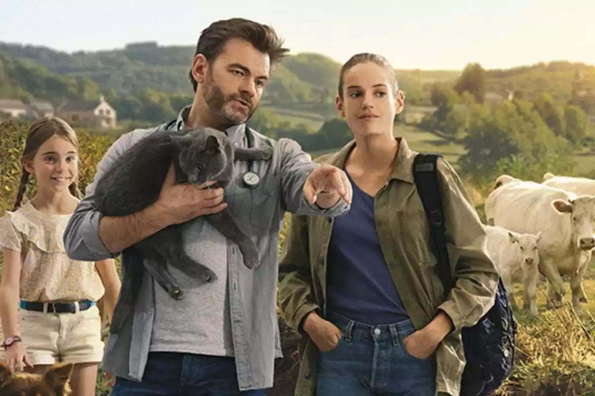 Home amb un gat els braços i noia en primer terme, amb nena i vaques al fons amb fons de natura