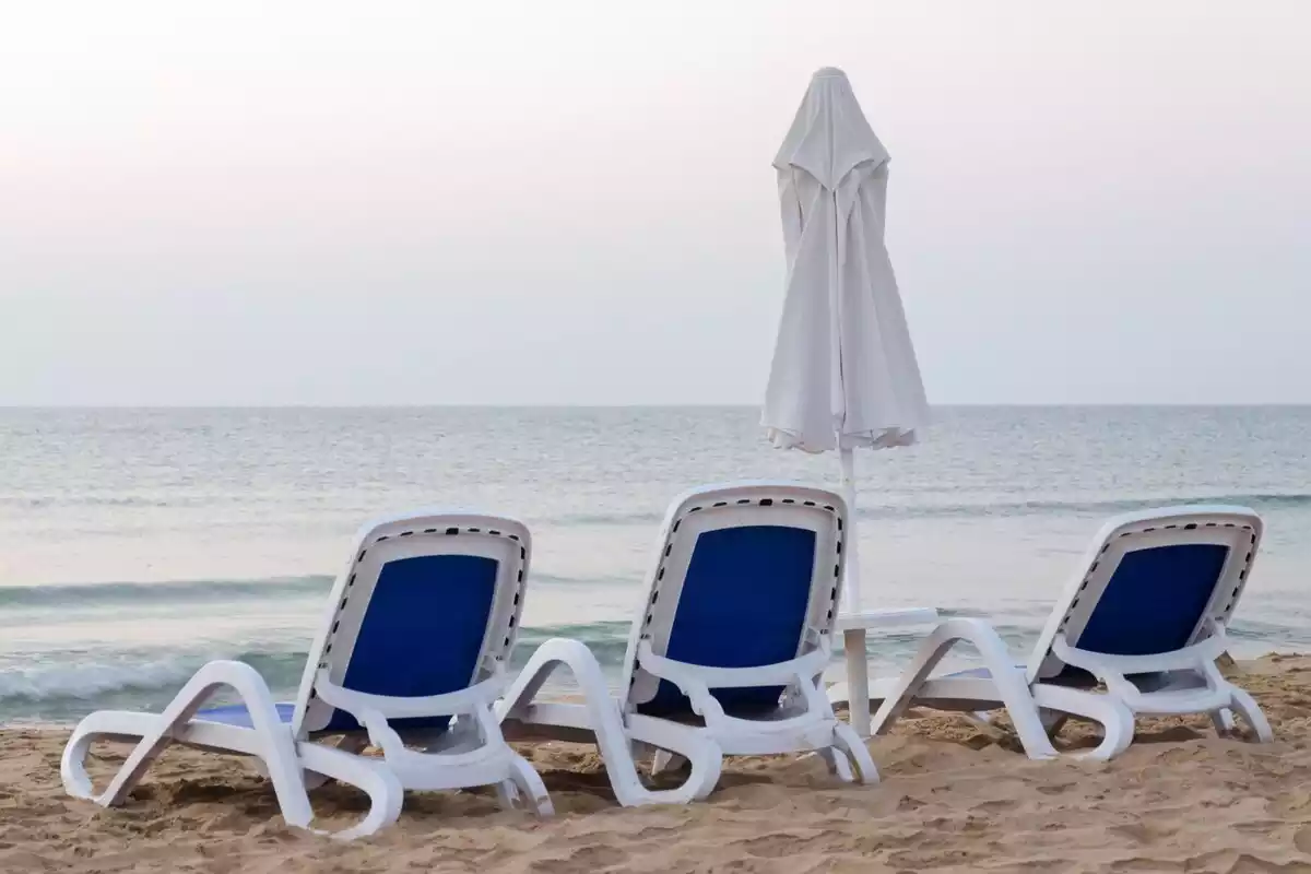 Imatge de tres cadires i un para-sol en una platja