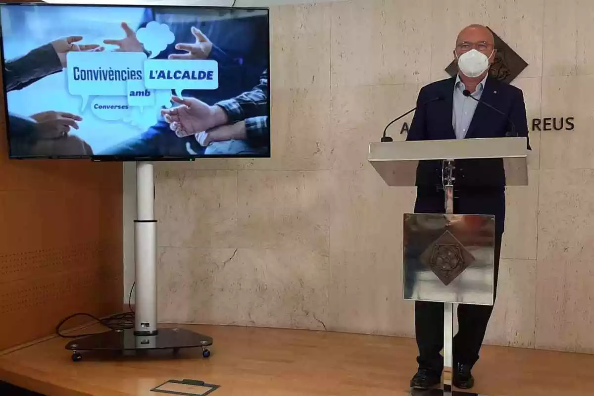 Carles Pellicer a la sala de premsa de l'Ajuntament de Reus amb una pantalla mostrant el programa de convivències als barris