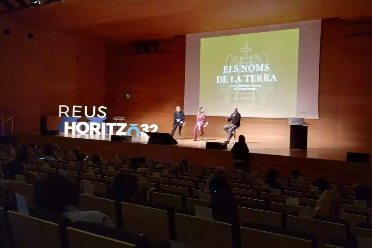 L'auditori de firaReus amb tres persones sobre l'escenari, una pantalla al darrere i el cartell 'Reus Horitzó 32'