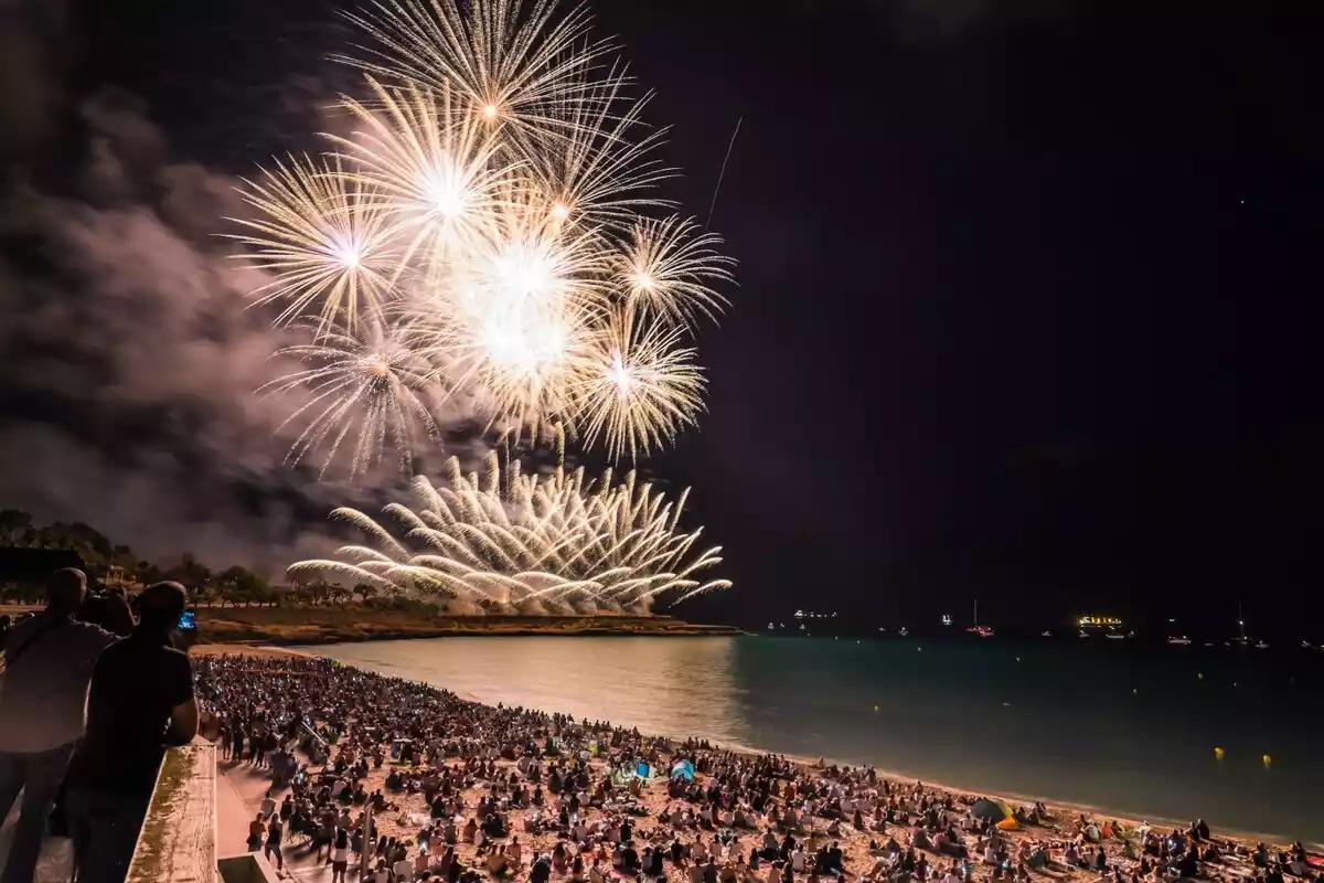 Imatge de la platja del Miracle de Tarragona plena de gent amb un castell de focs artificials al fons