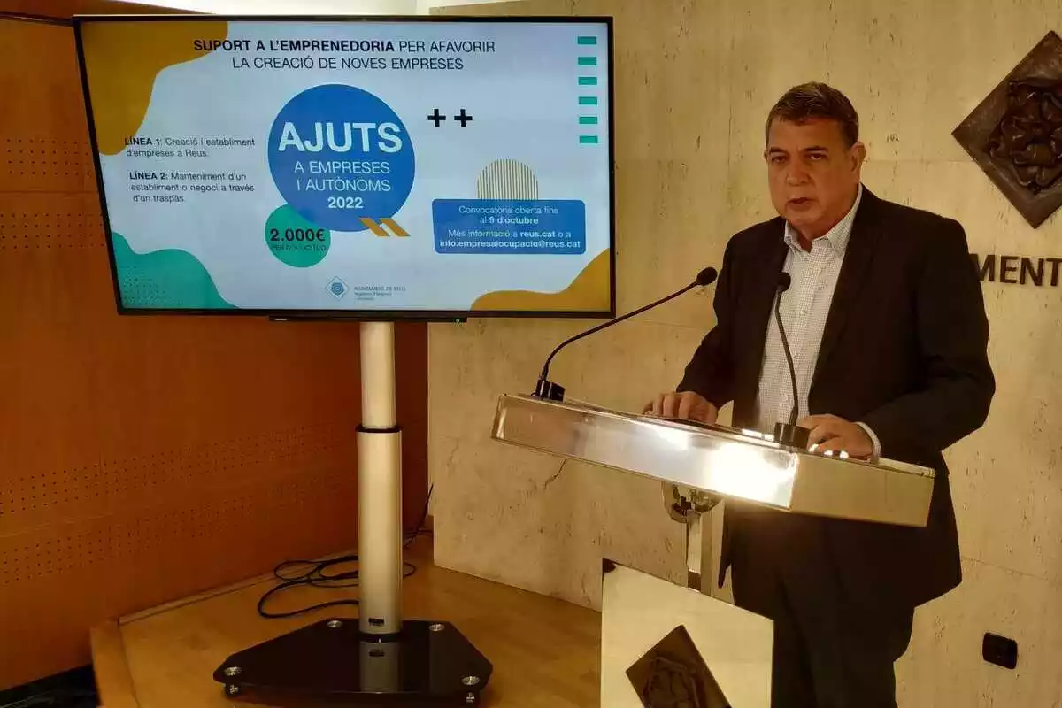 Carles Prats al faristol de la sala de premsa de l'Ajuntament de Reus, amb una pantalla al costat on es projecta la campanya d'ajuts a empreses