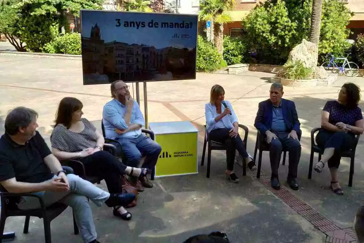 El grup municipal d'ERC Reus asseguts en semicercle a la plaça de Gabriel Ferraté, amb una pantalla amb el missatge «3 anys de mandat»