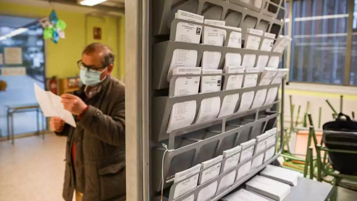 Imatge de paperetes electorals en un col·legi electoral, amb un home en segon pla preparant el seu vot