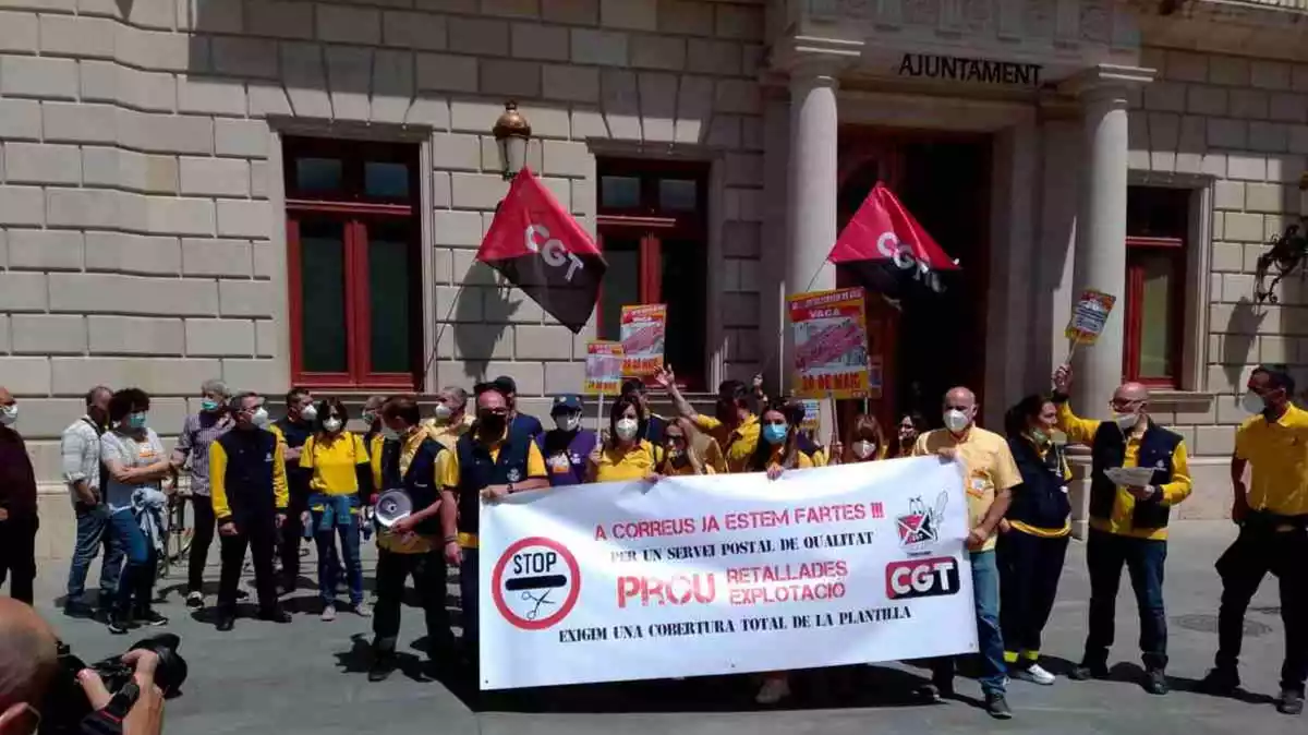 Una vintena llarga de persones, la majoria amb l'uniforme de Correus, davant de l'Ajuntament de Reus, amb una pancarta mostrant les seves reivindicacions