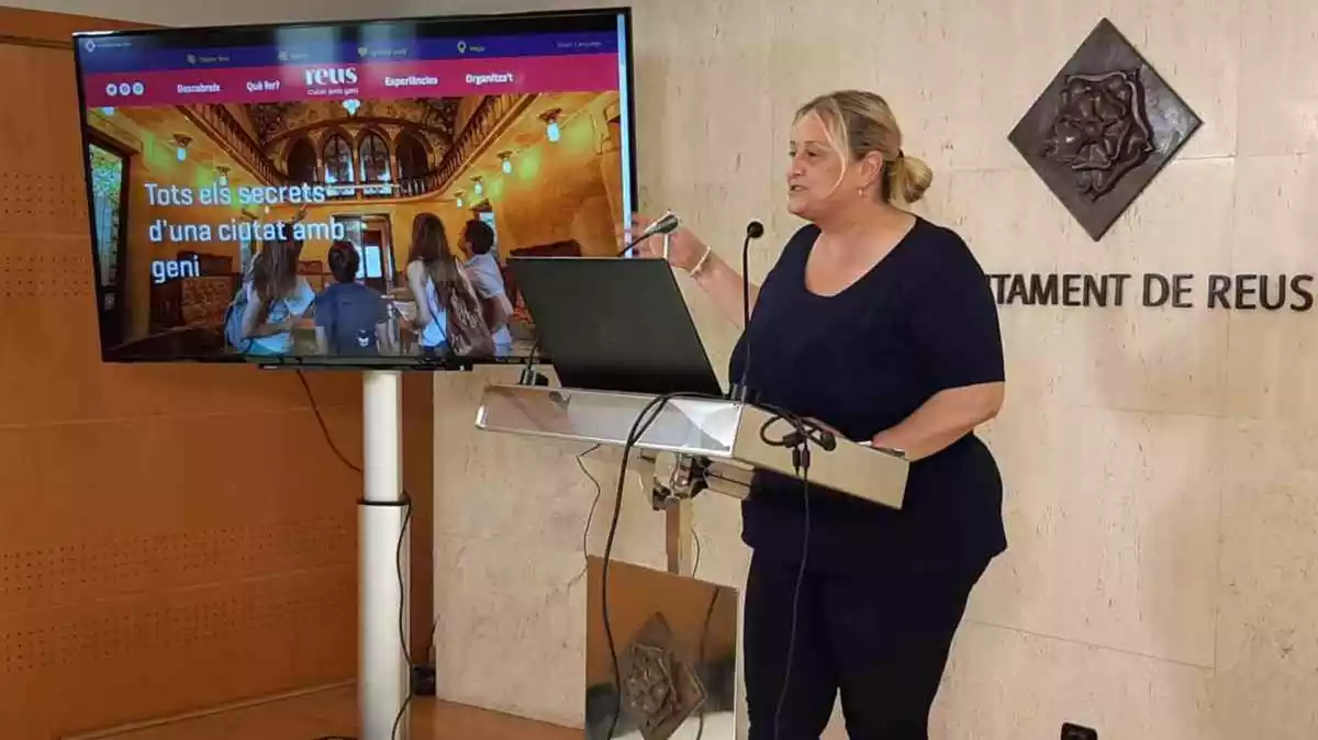 Montserrat Caelles a la tarima de la sala de premsa de l'Ajuntament de Reus amb una pantalla al costat