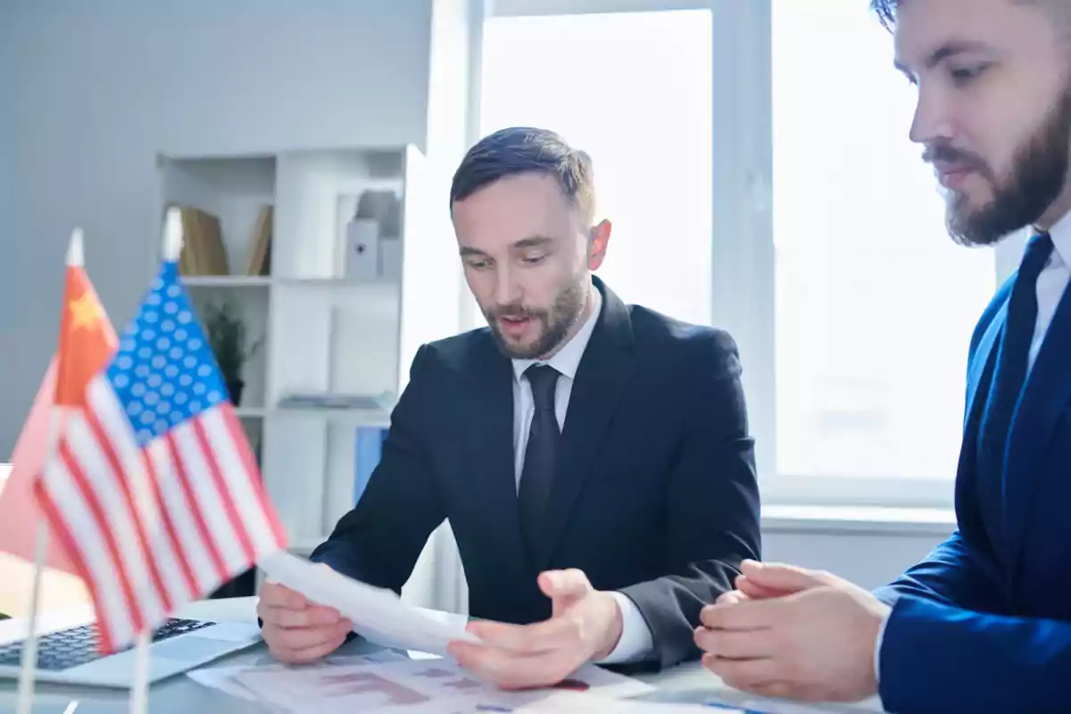 Dos hombres de negocios vestidos con traje revisan documentos en una oficina con banderas de Estados Unidos y China sobre la mesa