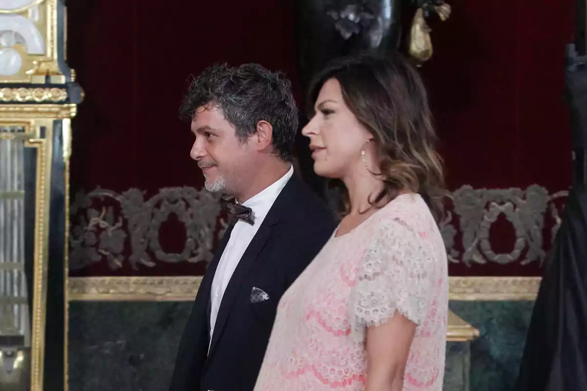 Alejandro Sanz amb esmòquing i Raquel Perera amb vestit clar caminen junts per un elegant saló decorat amb detalls daurats i parets vermelles. Alejandro Sanz amb esmòquing i Raquel Perera amb vestit clar caminen junts per un elegant saló decorat amb detalls daurats i parets vermelles.