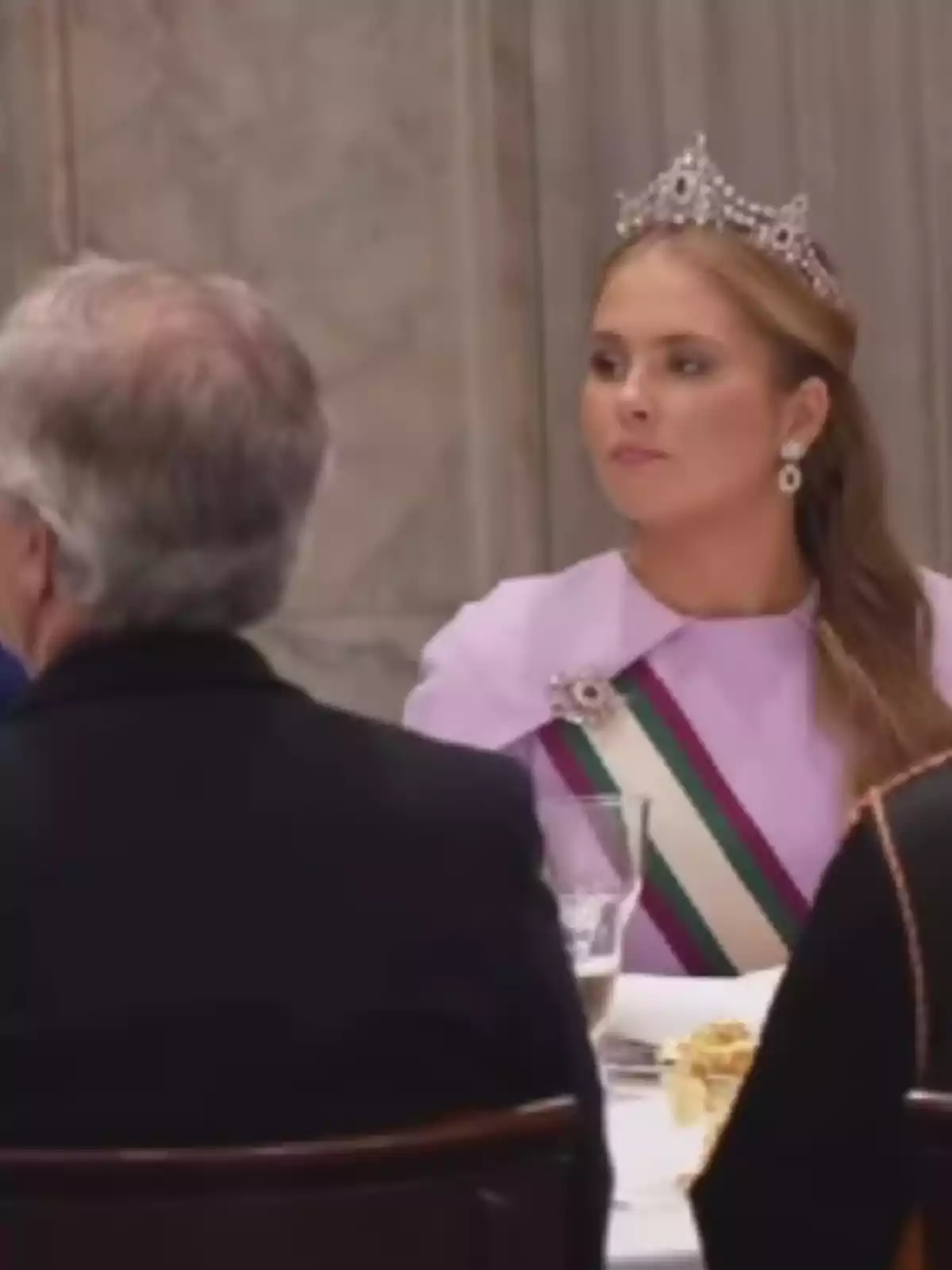 Dona amb tiara i banda cerimonial asseguda en una taula elegant durant un banquet Dona amb tiara i banda cerimonial asseguda en una taula elegant durant un banquet