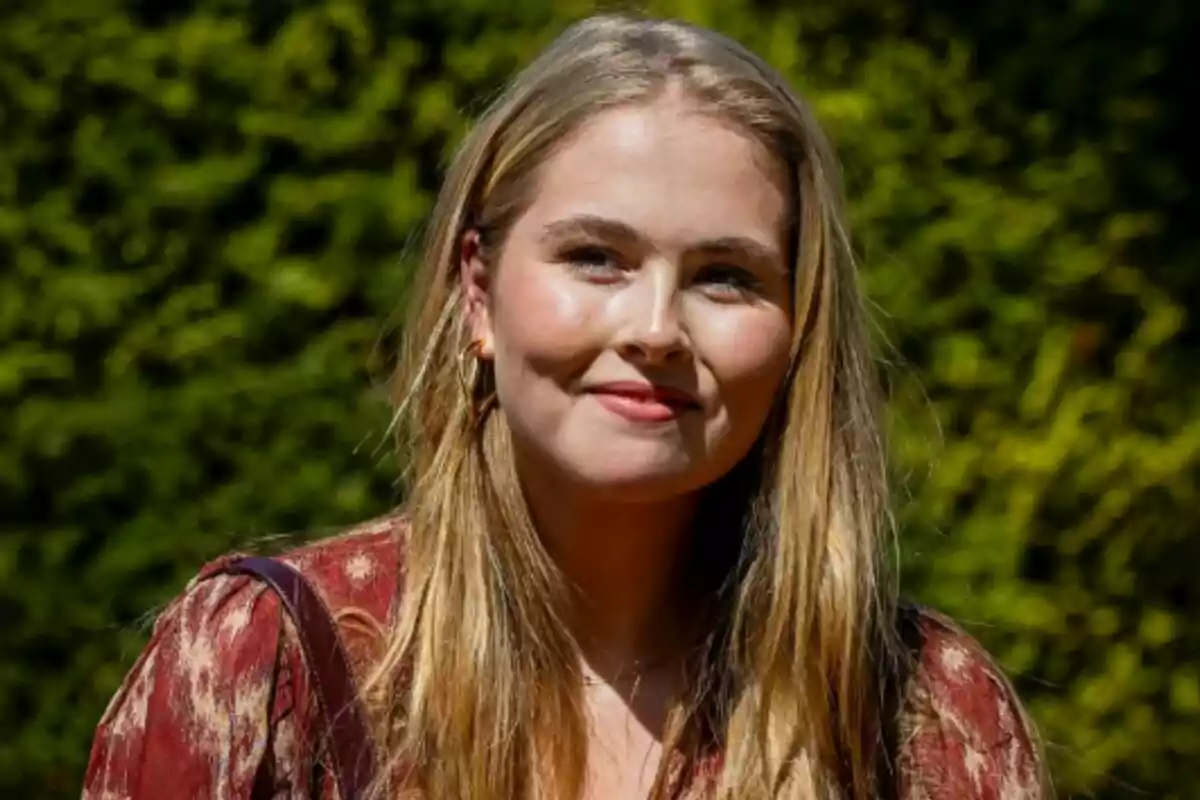 Close-up of Princess Amalia of the Netherlands wearing a patterned red blouse, smiling outdoors against a background of green vegetation. Close-up of Princess Amalia of the Netherlands wearing a patterned red blouse, smiling outdoors against a background of green vegetation.