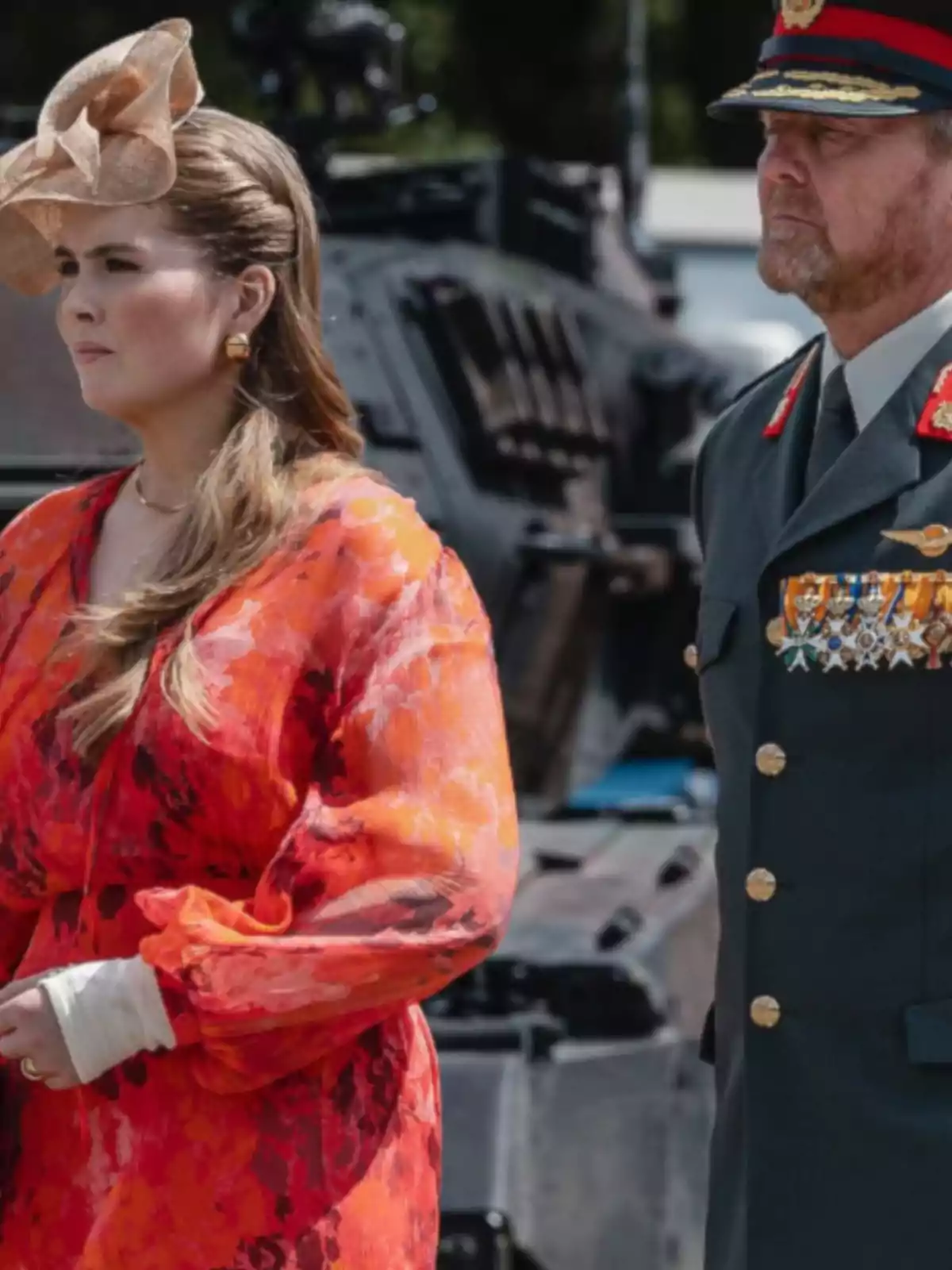 Una dona amb vestit vermell i tocat elegant és al costat d’un home amb uniforme militar i medalles, tots dos drets davant d’un vehicle blindat. Una dona amb vestit vermell i tocat elegant és al costat d’un home amb uniforme militar i medalles, tots dos drets davant d’un vehicle blindat.