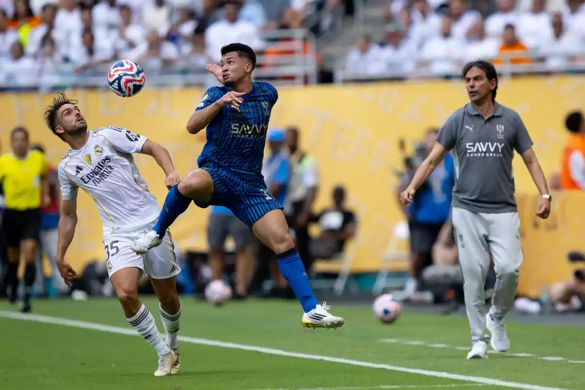 Dos futbolistas disputan el balón en el aire durante un partido mientras un entrenador observa desde la línea de banda Dos futbolistas disputan el balón en el aire durante un partido mientras un entrenador observa desde la línea de banda