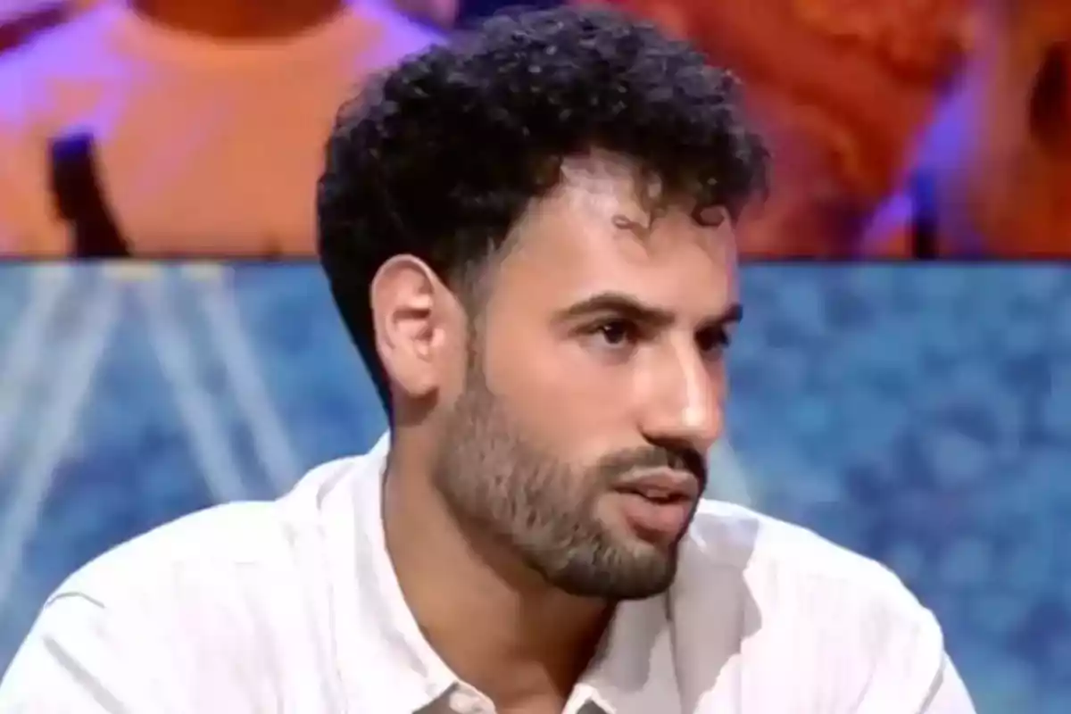 Asraf Beno de cabello rizado y barba corta con camisa clara participando en un programa de televisión Asraf Beno de cabello rizado y barba corta con camisa clara participando en un programa de televisión
