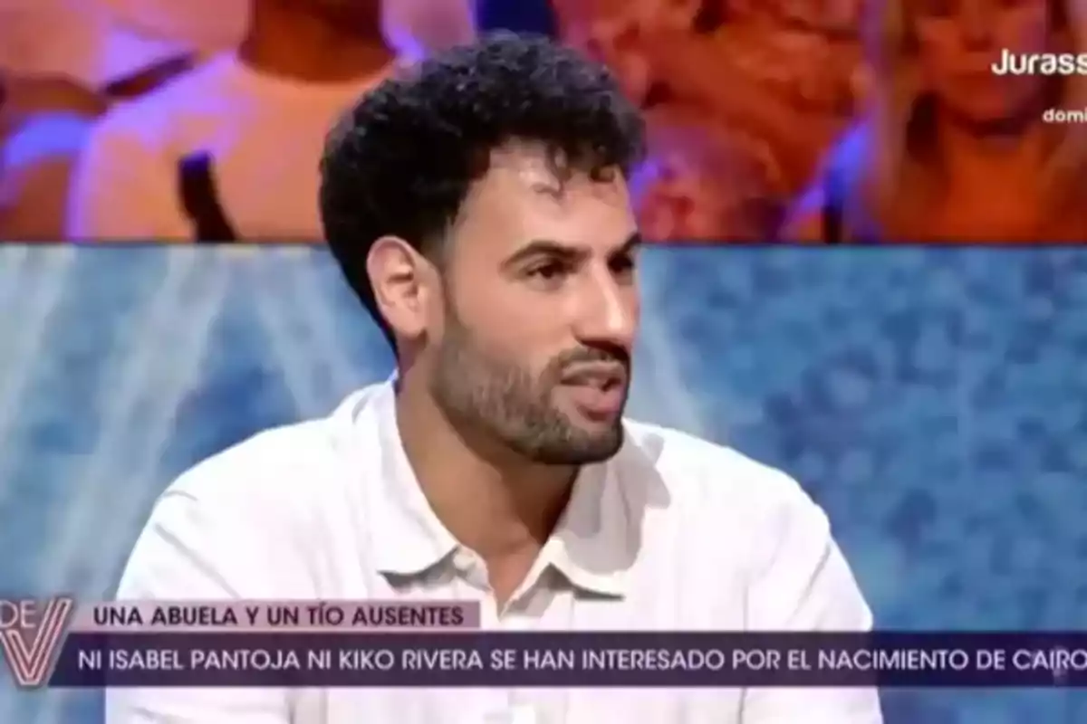 Asraf Beno de cabello rizado y barba corta vestido con camisa clara participando en un programa de televisión con subtítulos en la parte inferior de la pantalla. Asraf Beno de cabello rizado y barba corta vestido con camisa clara participando en un programa de televisión con subtítulos en la parte inferior de la pantalla.