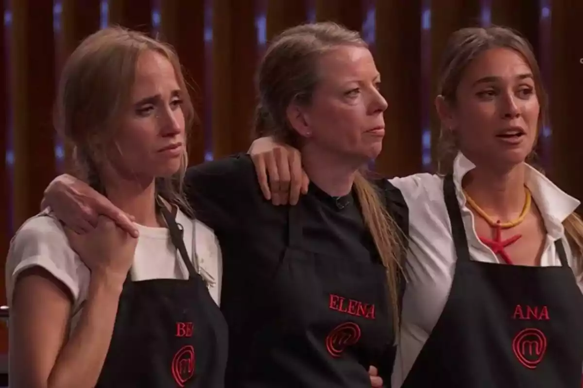 Tres dones –Bea, Elena i Ana– amb davantals negres de MasterChef s’abracen i mostren expressions d’emoció i nerviosisme en un plató de cuina il·luminat. Tres dones –Bea, Elena i Ana– amb davantals negres de MasterChef s’abracen i mostren expressions d’emoció i nerviosisme en un plató de cuina il·luminat.