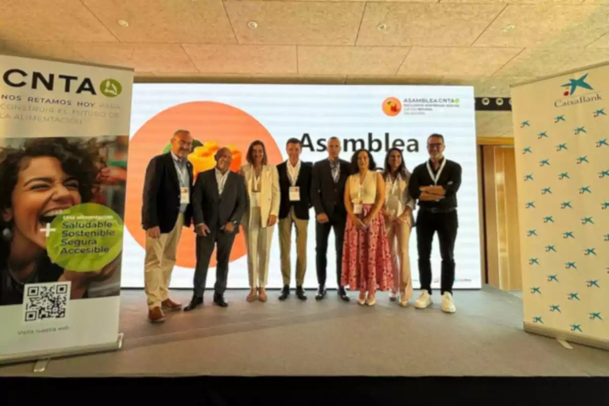 Un grup de persones posant en un escenari durant un esdeveniment corporatiu amb pancartes de CNTA i CaixaBank als costats i una pantalla gran de fons que mostra la paraula Assemblea Un grup de persones posant en un escenari durant un esdeveniment corporatiu amb pancartes de CNTA i CaixaBank als costats i una pantalla gran de fons que mostra la paraula Assemblea