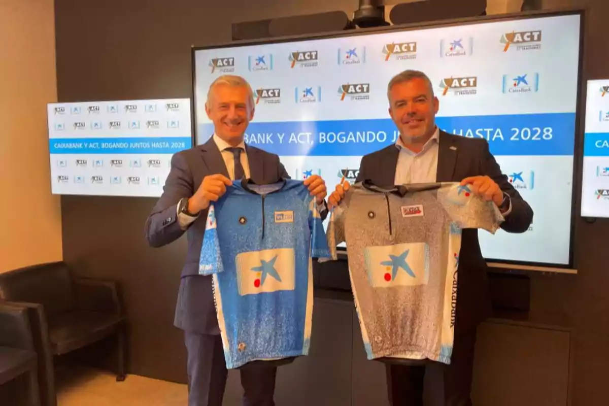Dos homes amb vestit somriuen mentre sostenen samarretes esportives davant d’una pantalla amb logotips de Caixabank i ACT Dos homes amb vestit somriuen mentre sostenen samarretes esportives davant d’una pantalla amb logotips de Caixabank i ACT