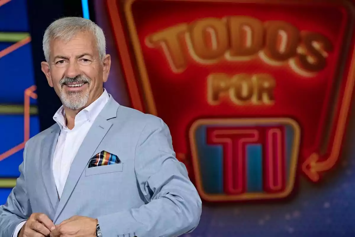 Carlos Sobera, de cabell canós i barba, vestit amb americana blau clar i camisa blanca, somriu davant d’un rètol colorit que diu Carlos Sobera, de cabell canós i barba, vestit amb americana blau clar i camisa blanca, somriu davant d’un rètol colorit que diu