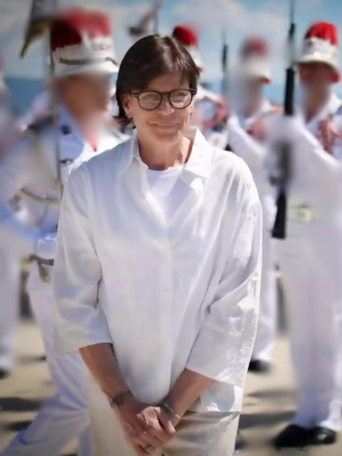 Dona de cabell curt i ulleres vestida de blanc somrient a l’aire lliure amb persones uniformades i desenfocades al fons Dona de cabell curt i ulleres vestida de blanc somrient a l’aire lliure amb persones uniformades i desenfocades al fons