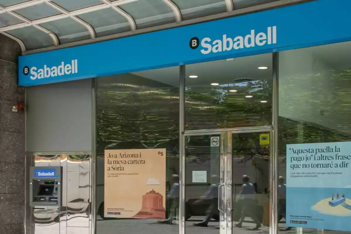 Façana d’una sucursal del banc Sabadell amb un caixer automàtic i cartells publicitaris a les finestres. Façana d’una sucursal del banc Sabadell amb un caixer automàtic i cartells publicitaris a les finestres.