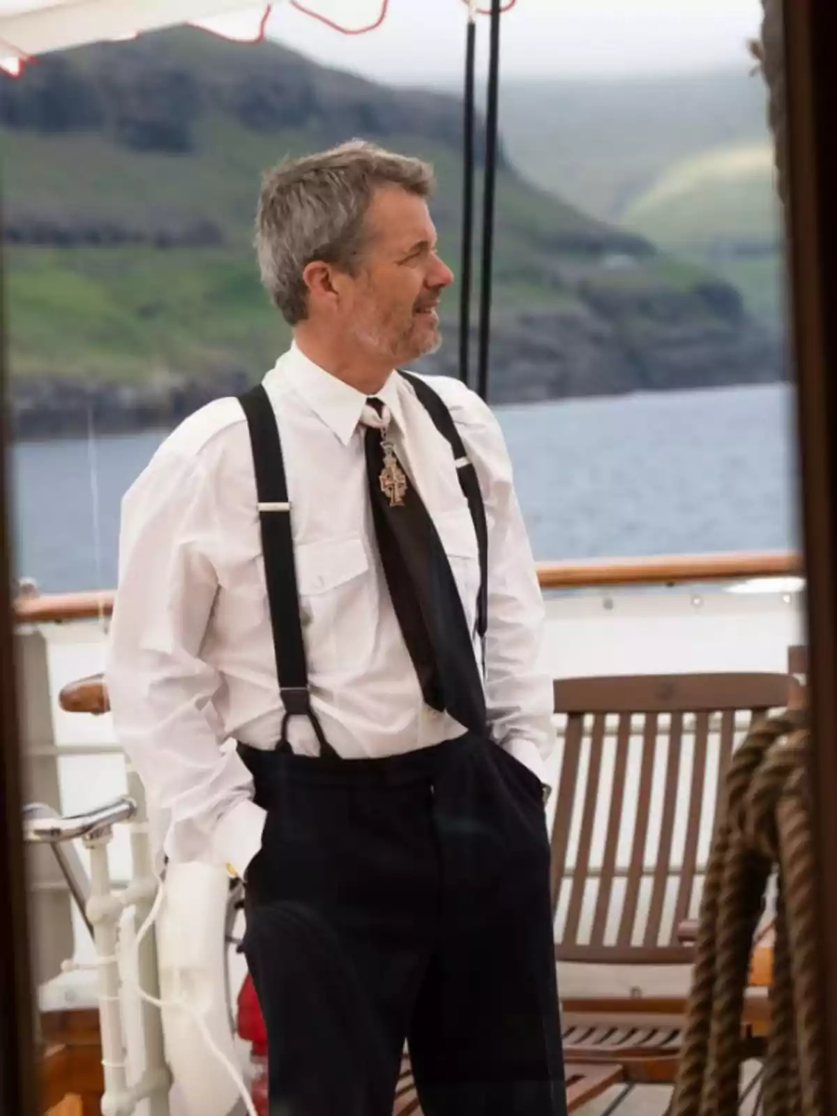 Home de cabells canosos i barba vestit amb camisa blanca, tirants i corbata negra, dret a la coberta d’un vaixell amb un paisatge de muntanyes i aigua al fons Home de cabells canosos i barba vestit amb camisa blanca, tirants i corbata negra, dret a la coberta d’un vaixell amb un paisatge de muntanyes i aigua al fons