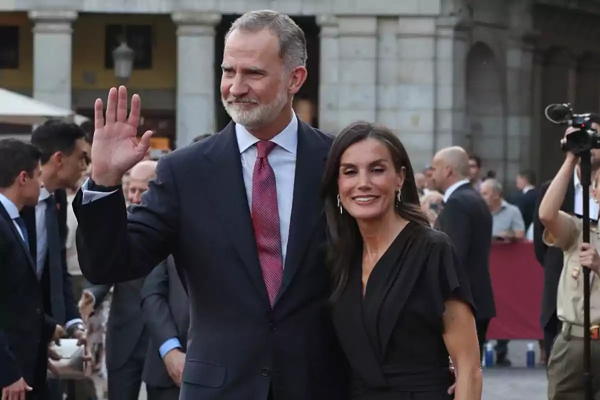 Felipe VI in a dark suit and red tie waves while Queen Letizia, wearing a black dress, smiles beside him at an outdoor event surrounded by other people. Felipe VI in a dark suit and red tie waves while Queen Letizia, wearing a black dress, smiles beside him at an outdoor event surrounded by other people.