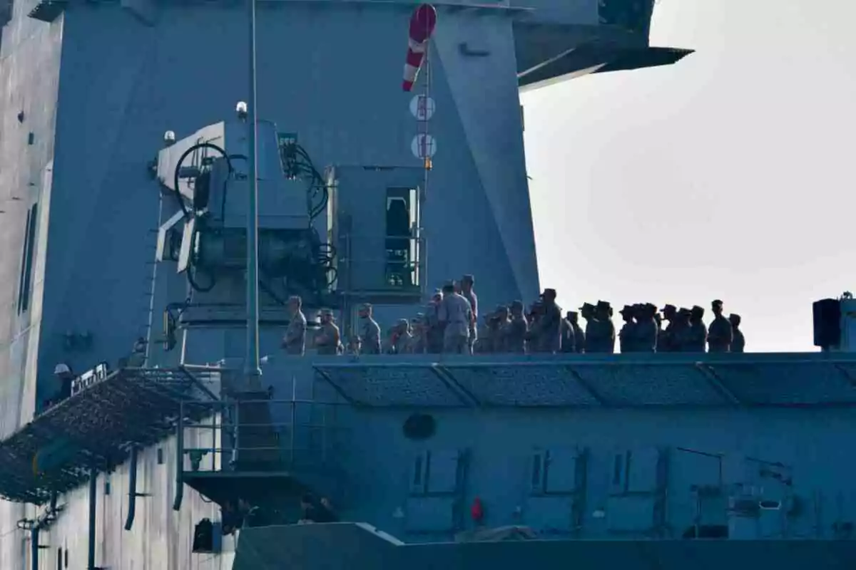 Un grupo de militares uniformados se encuentra de pie sobre la cubierta de un buque gris, observando hacia el exterior. Un grupo de militares uniformados se encuentra de pie sobre la cubierta de un buque gris, observando hacia el exterior.