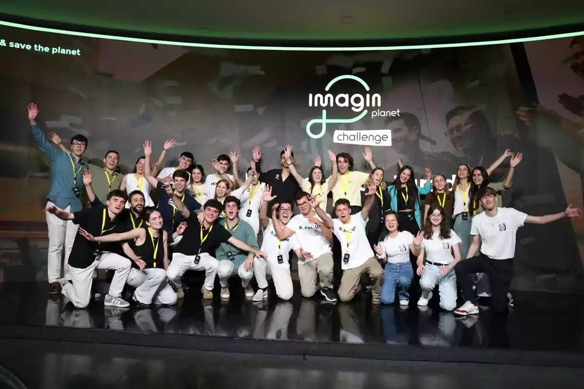 Grup de joves somrients posant junts en un escenari amb samarretes i credencials, aixecant els braços davant d’un fons que diu imagin planet challenge. Grup de joves somrients posant junts en un escenari amb samarretes i credencials, aixecant els braços davant d’un fons que diu imagin planet challenge.