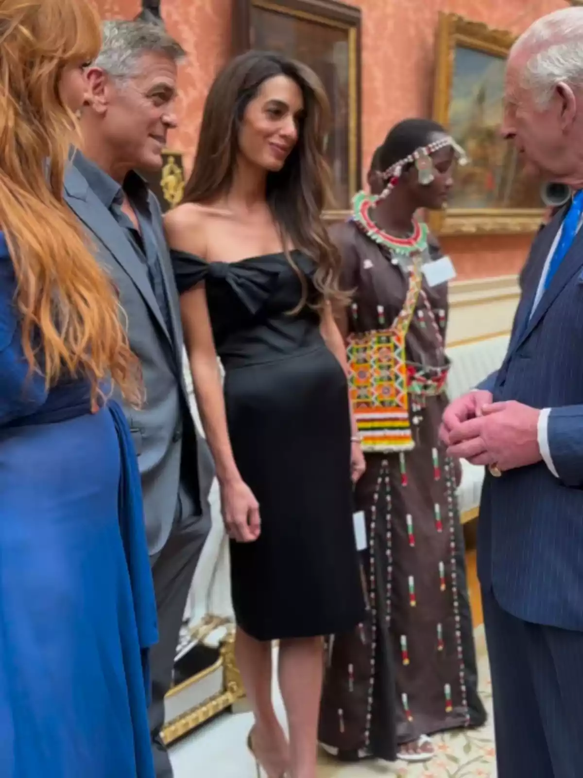 Diverses persones elegantment vestides conversen en una sala decorada amb quadres i mobles clàssics, una d’elles porta un vestit tradicional amb comptes de colors. Diverses persones elegantment vestides conversen en una sala decorada amb quadres i mobles clàssics, una d’elles porta un vestit tradicional amb comptes de colors.
