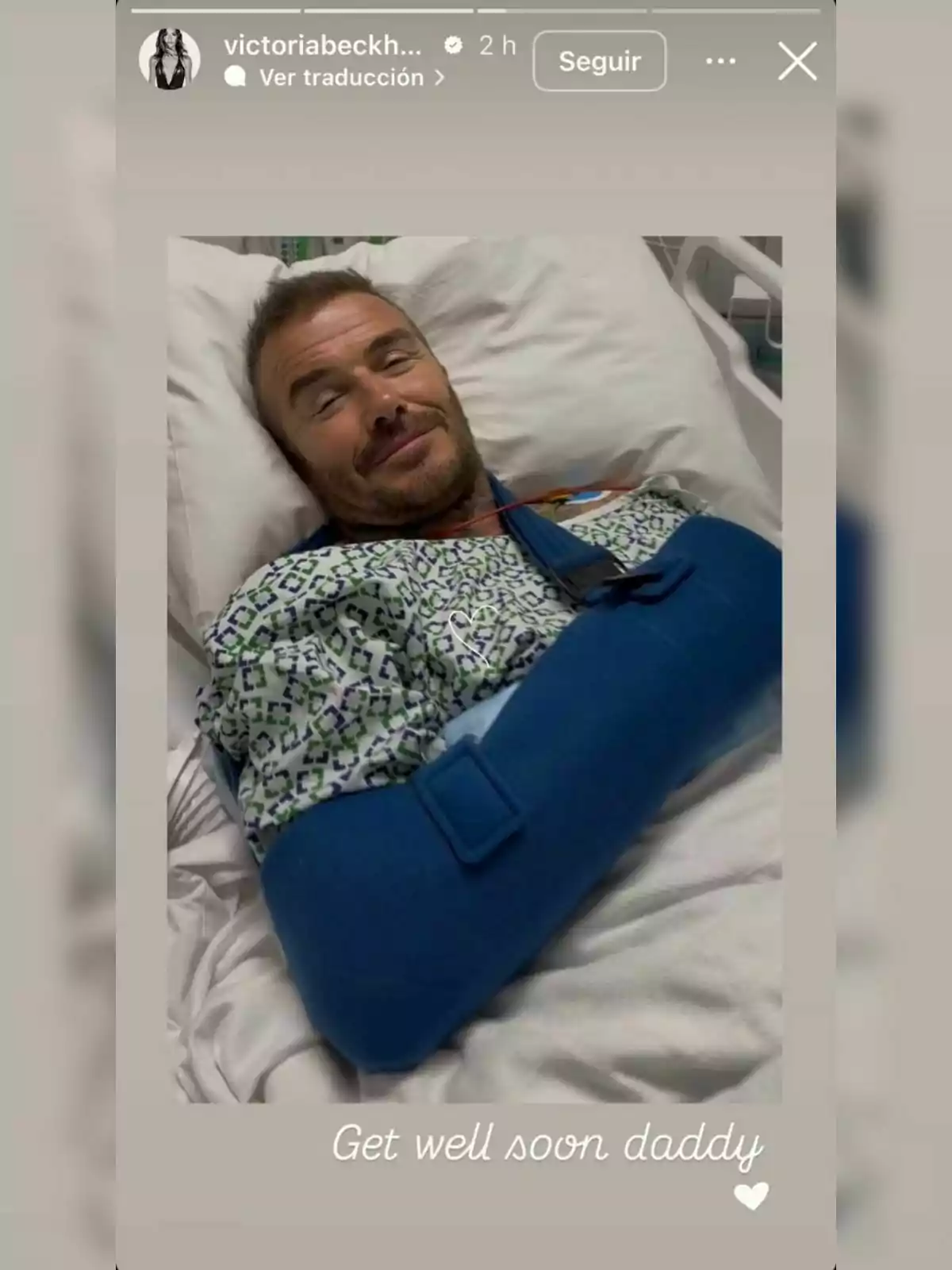 David Beckham lying on a hospital bed wearing a medical gown and a blue sling on his left arm, accompanied by the text Get well soon daddy and a white heart. David Beckham lying on a hospital bed wearing a medical gown and a blue sling on his left arm, accompanied by the text Get well soon daddy and a white heart.