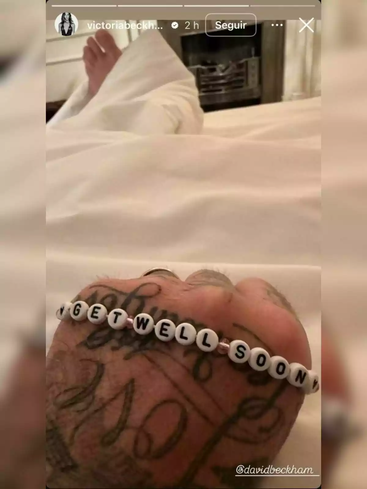 Tattooed hand of David Beckham holding a beaded bracelet that says GET WELL SOON over a white bed, with a foot covered by a sheet in the background. Tattooed hand of David Beckham holding a beaded bracelet that says GET WELL SOON over a white bed, with a foot covered by a sheet in the background.