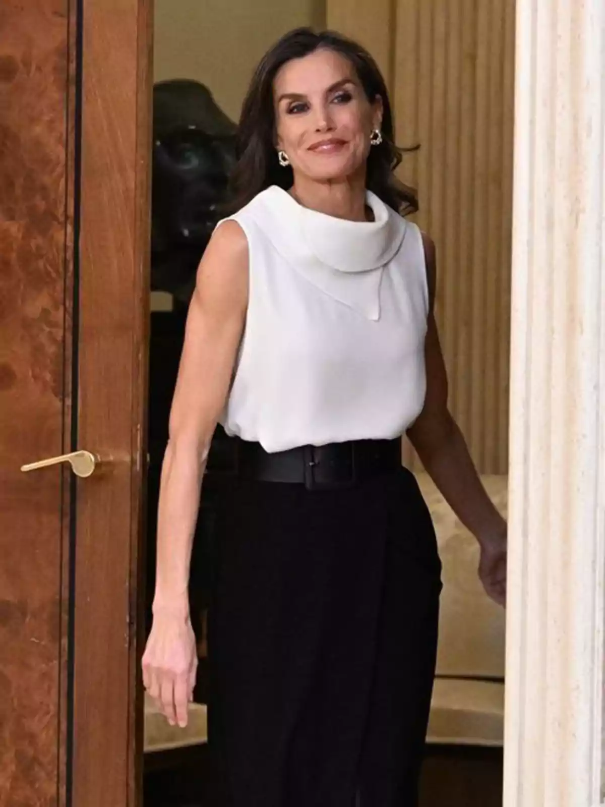 Dona de cabell fosc i solt entrant per una porta, vestida amb una brusa blanca de tirants i uns pantalons negres de cintura alta amb cinturó Dona de cabell fosc i solt entrant per una porta, vestida amb una brusa blanca de tirants i uns pantalons negres de cintura alta amb cinturó