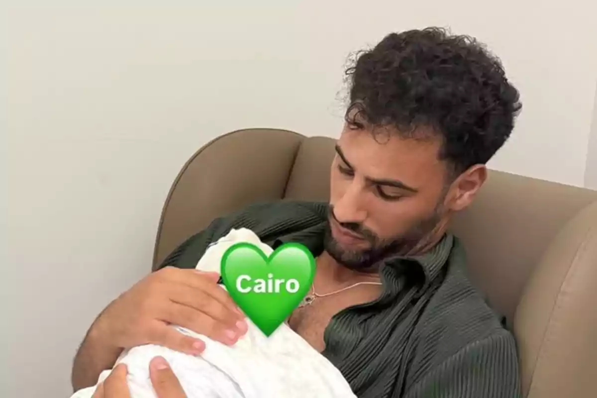 Home assegut en un sofà sostenint un nadó embolicat amb una manta blanca amb un emoji de cor verd que diu Cairo cobrint la cara del nadó Home assegut en un sofà sostenint un nadó embolicat amb una manta blanca amb un emoji de cor verd que diu Cairo cobrint la cara del nadó