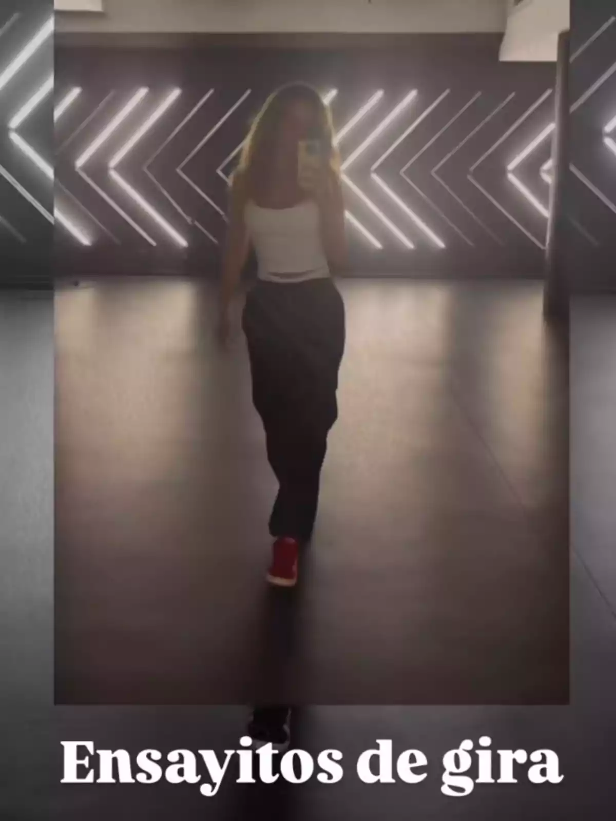 Persona fent-se una selfie davant d’un mirall en un estudi de dansa amb llums a la paret i el text Assajos de gira a la part inferior Persona fent-se una selfie davant d’un mirall en un estudi de dansa amb llums a la paret i el text Assajos de gira a la part inferior