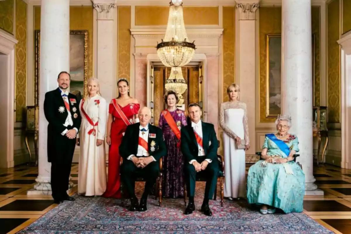 Ocho personas elegantemente vestidas posan en un salón lujoso con columnas blancas, alfombra decorativa y una gran lámpara de araña al fondo Ocho personas elegantemente vestidas posan en un salón lujoso con columnas blancas, alfombra decorativa y una gran lámpara de araña al fondo