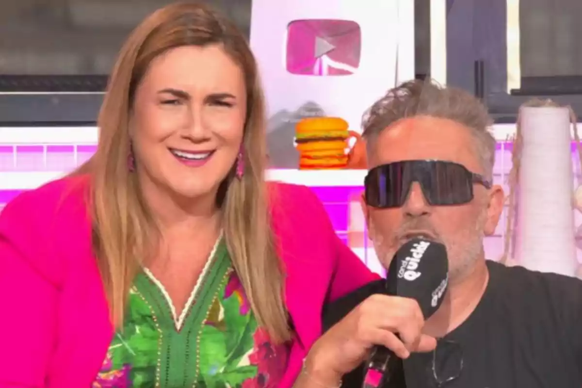 Dues persones en un plató de gravació, una dona somrient amb jaqueta rosa i un home amb ulleres de sol sostenint un micròfon, al fons un botó de YouTube platejat i decoració colorida. Dues persones en un plató de gravació, una dona somrient amb jaqueta rosa i un home amb ulleres de sol sostenint un micròfon, al fons un botó de YouTube platejat i decoració colorida.