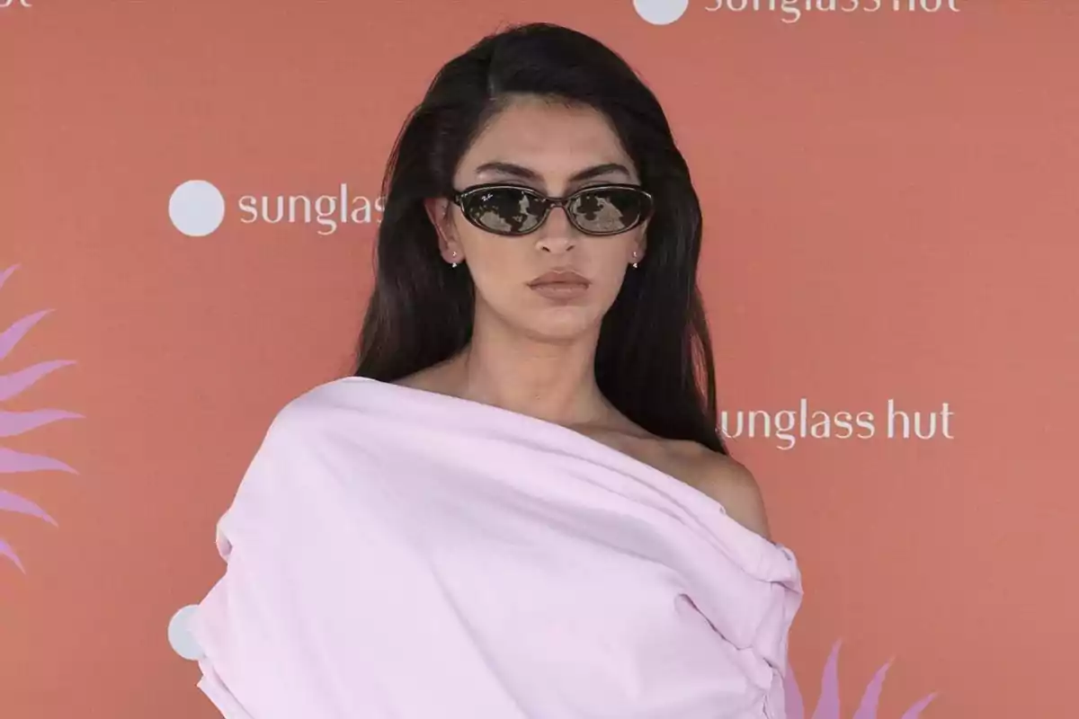Woman with long dark hair wearing sunglasses and a light pink dress posing in front of an orange background with a glasses store logo Woman with long dark hair wearing sunglasses and a light pink dress posing in front of an orange background with a glasses store logo