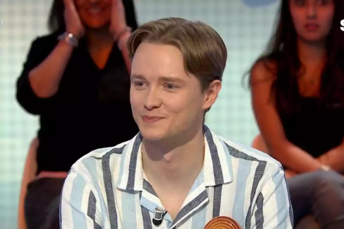 Jove de cabells rossos i camisa de ratlles somriu mentre participa en un programa de televisió amb públic al fons Jove de cabells rossos i camisa de ratlles somriu mentre participa en un programa de televisió amb públic al fons